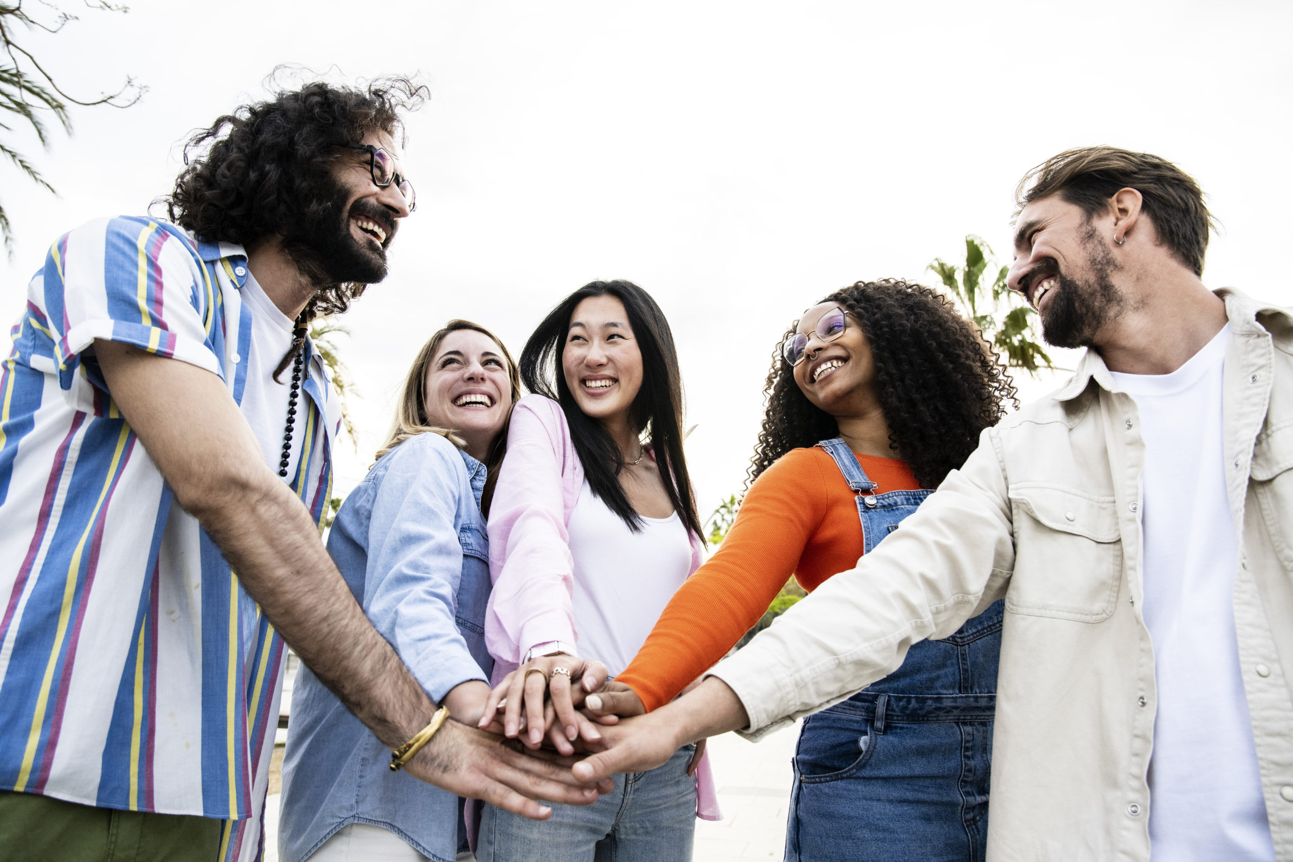 Diverse group of cheerful millennial friends standing together stacking hands in the street. Cheerful multiethnic young carefree friends laughing and having fun outside joining hands on the center.