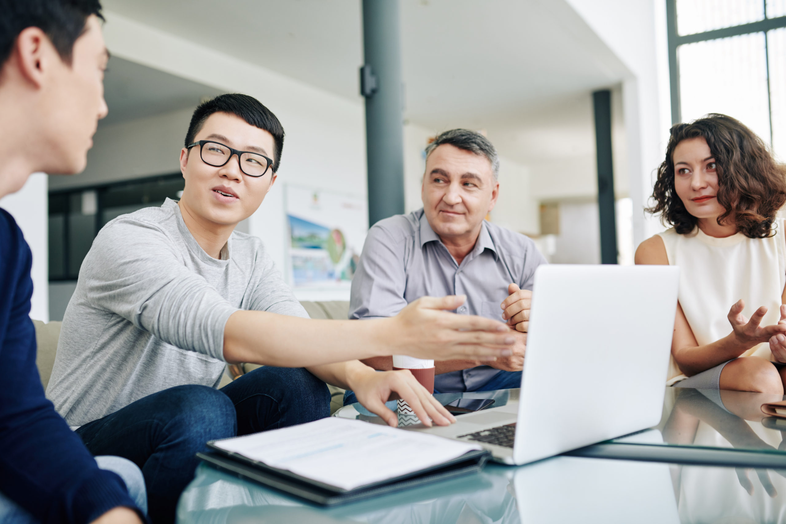 Smiling young Asian entrepreneur pointing at laptop screen when discussing figures in financial report with colleagues at meeting