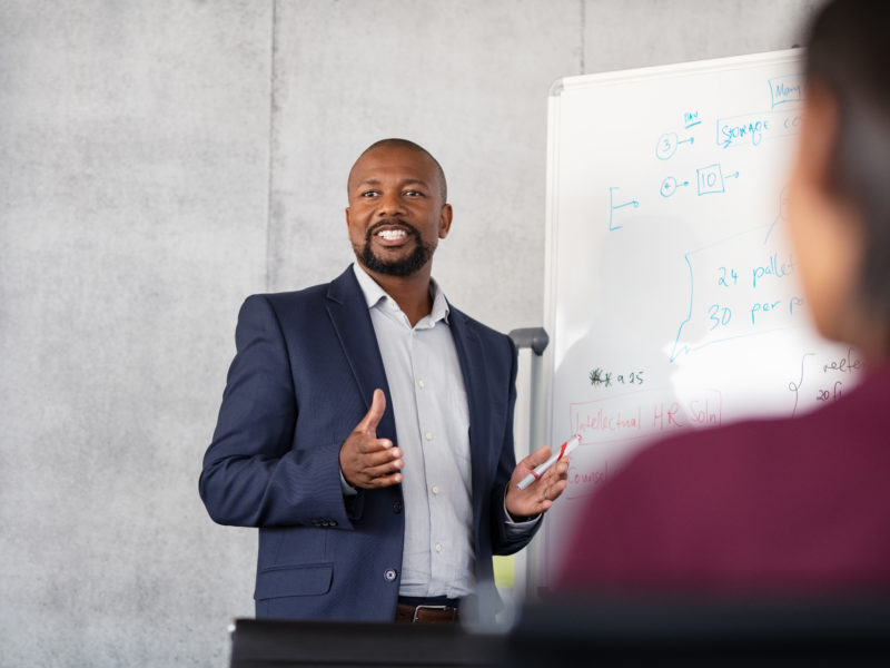 Mature african american coach explaining strategy to his team with copy space. Successful businessman presenting new project to employees during meeting room. Conference speaker talk to audience while giving presentation on whiteboard to business group of people.