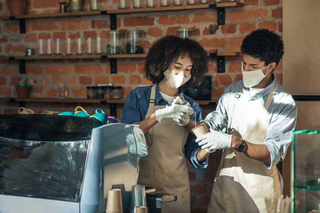 New employee, training and work during covid-19 pandemic. Millennial african american woman in apron, protective mask and gloves with guy preparing latte near equipment in interior of loft cafe