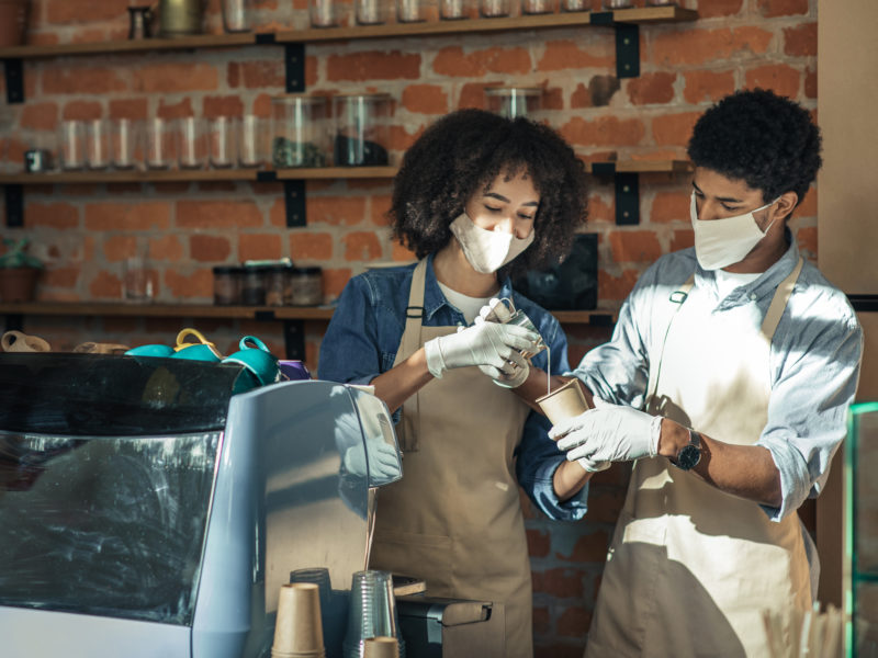 New employee, training and work during covid-19 pandemic. Millennial african american woman in apron, protective mask and gloves with guy preparing latte near equipment in interior of loft cafe