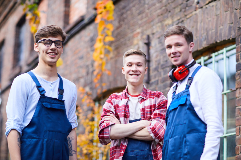 Group of cheerful young workers in uniforms standing against brick building outdoors and smiling at camera