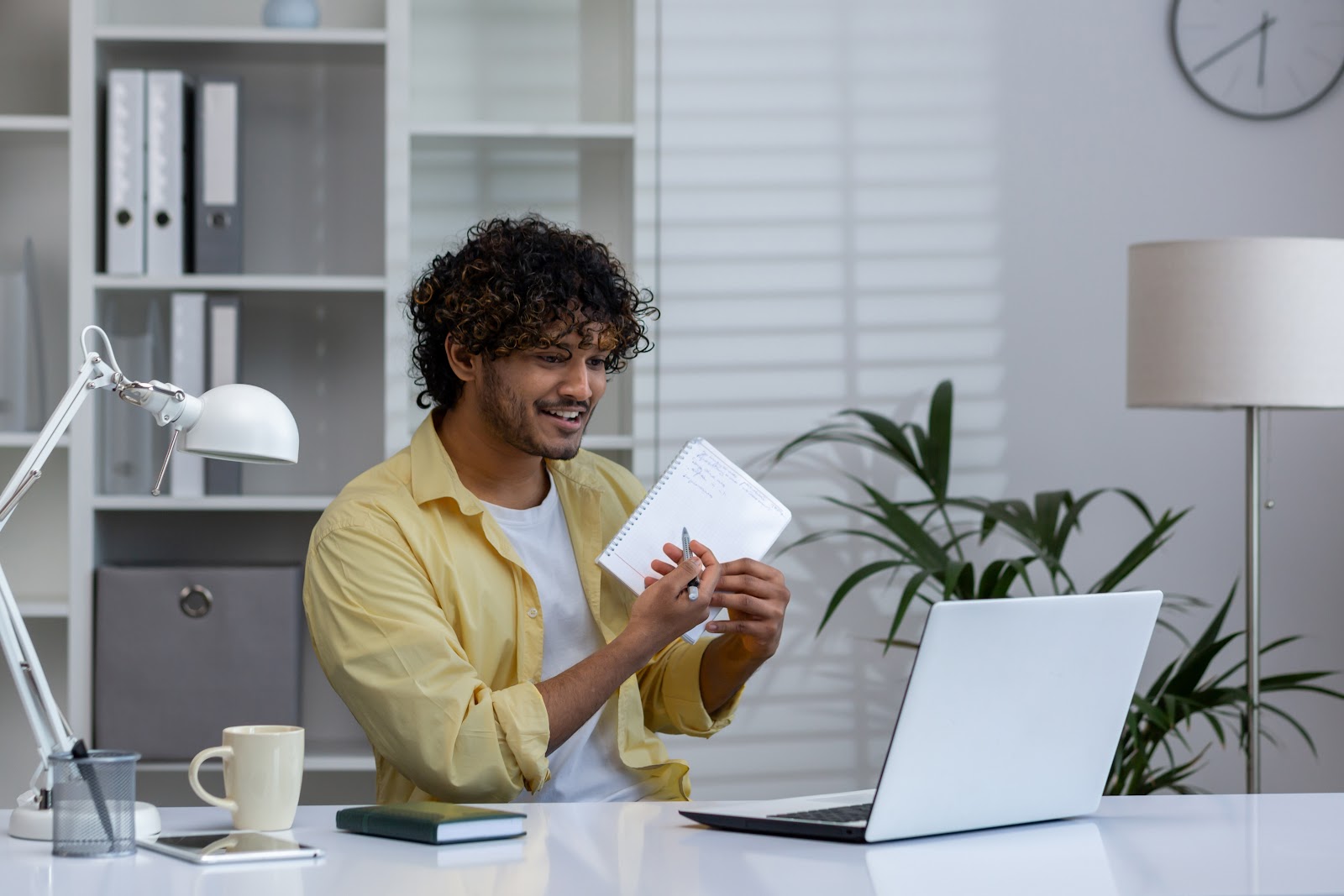 Smiling man working from home, holding a notebook and engaging in a virtual meeting from his home office. Remote work concept.