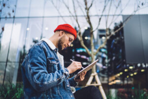Man sitting on street against glass skyscraper using a tablet with stylus