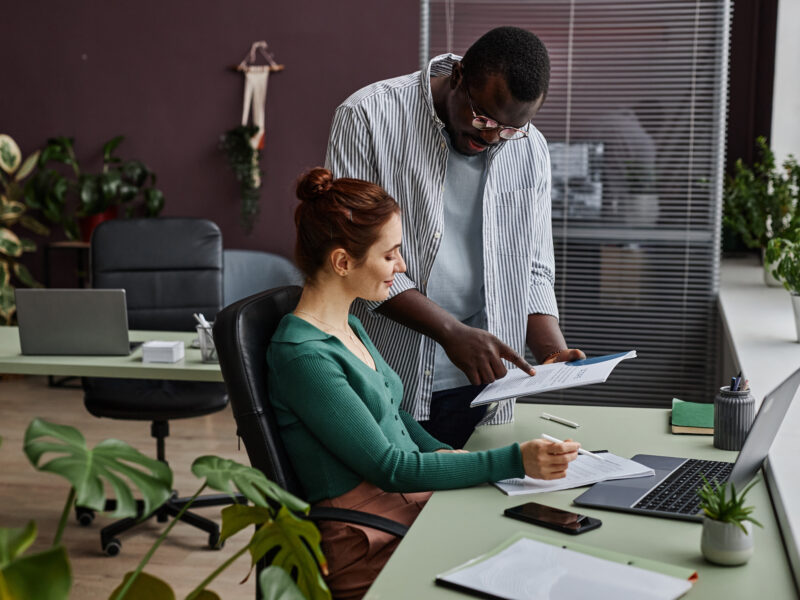Side view portrait of two people discussing project and reading documents at workplace in office decorated with plants