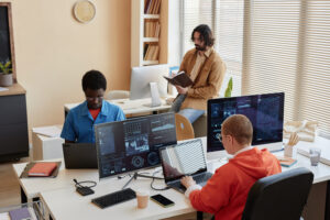 Group of young coworkers analyzing data while sitting in front of computers while one of them looking through notes in notebook