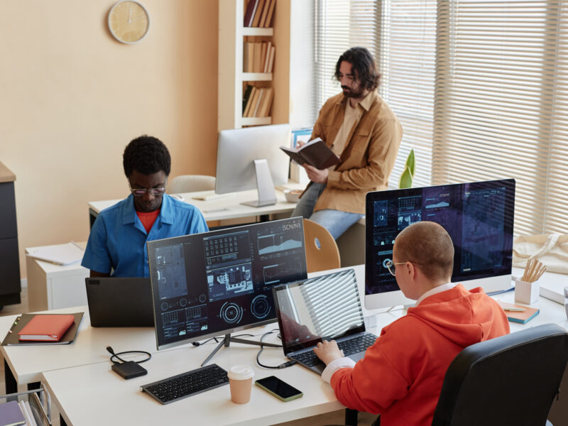 Group of young coworkers analyzing data while sitting in front of computers while one of them looking through notes in notebook
