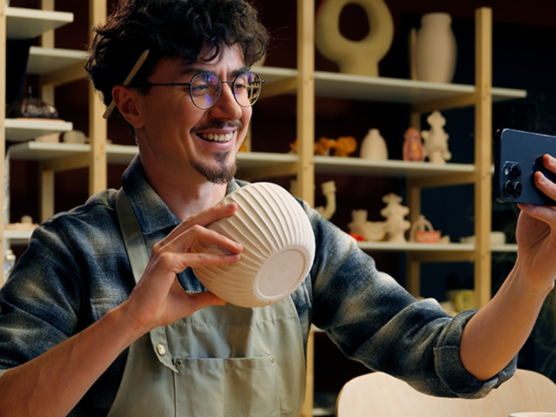 Smiling artisan in a pottery studio holding a ceramic bowl and filming it with a smartphone.