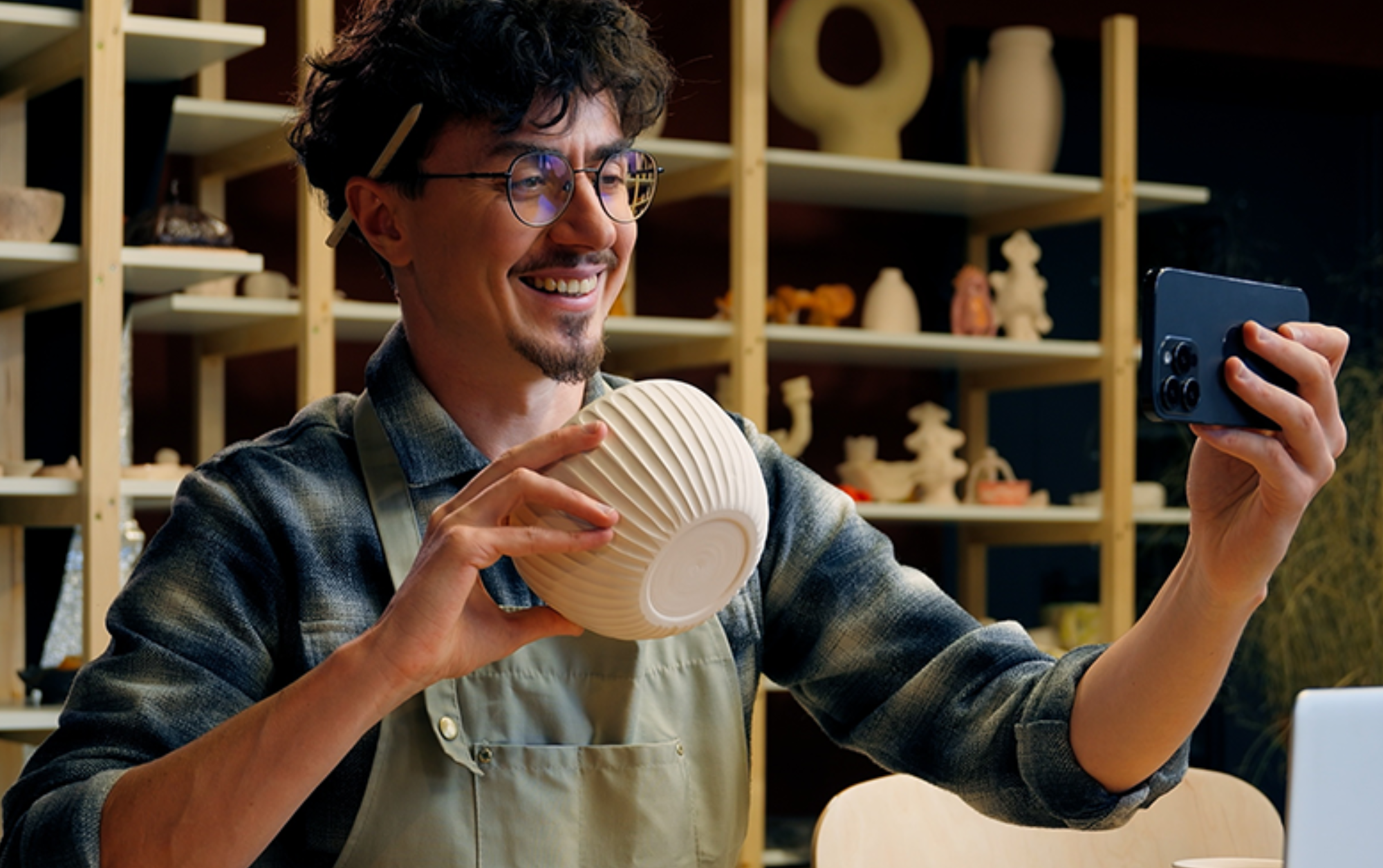 Smiling artisan in a pottery studio holding a ceramic bowl and filming it with a smartphone.