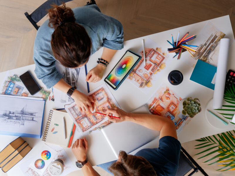 Two people collaborating on interior design sketches at a desk with color swatches and drawings.