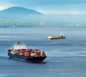 Cargo ships carrying stacked containers across calm water with a bridge and mountains in the background.