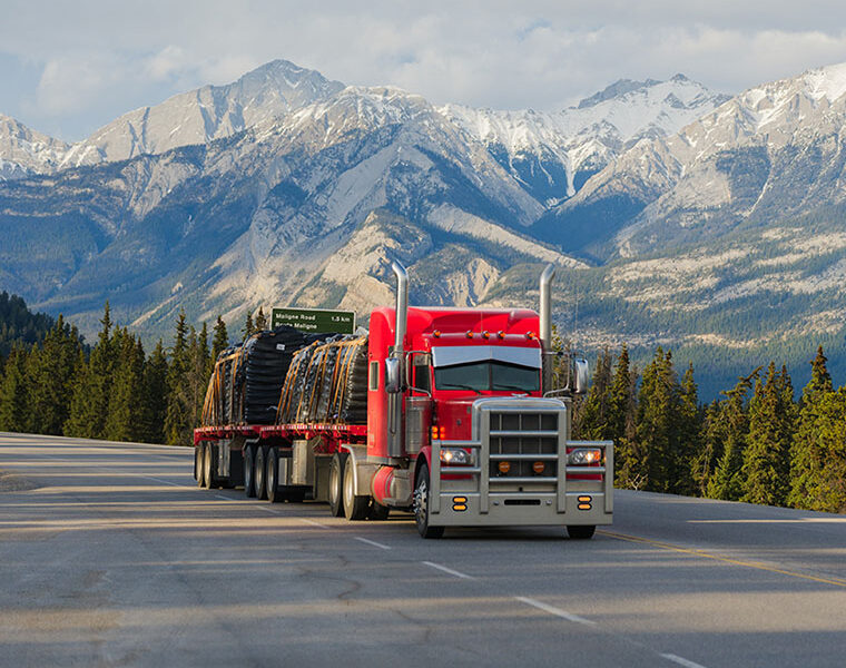 A red semi-truck hauling freight on a mountain highway, with snow-capped Rocky Mountain peaks and evergreen forest in the background.