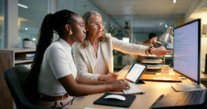 Two colleagues at a desk reviewing content on a computer, with one pointing at the screen while the other types.