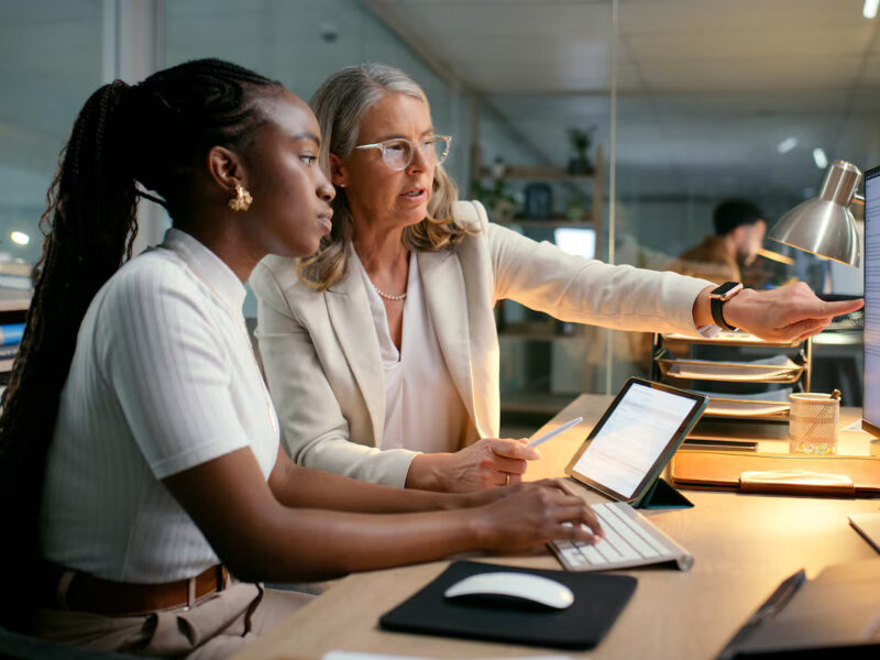 Two colleagues at a desk reviewing content on a computer, with one pointing at the screen while the other types.