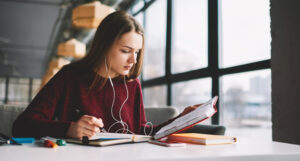 Pensive hipster girl learning language online via earphones using application while writing new words into textbook, attractive smart woman enjoying playlist music at cafeteria and studying