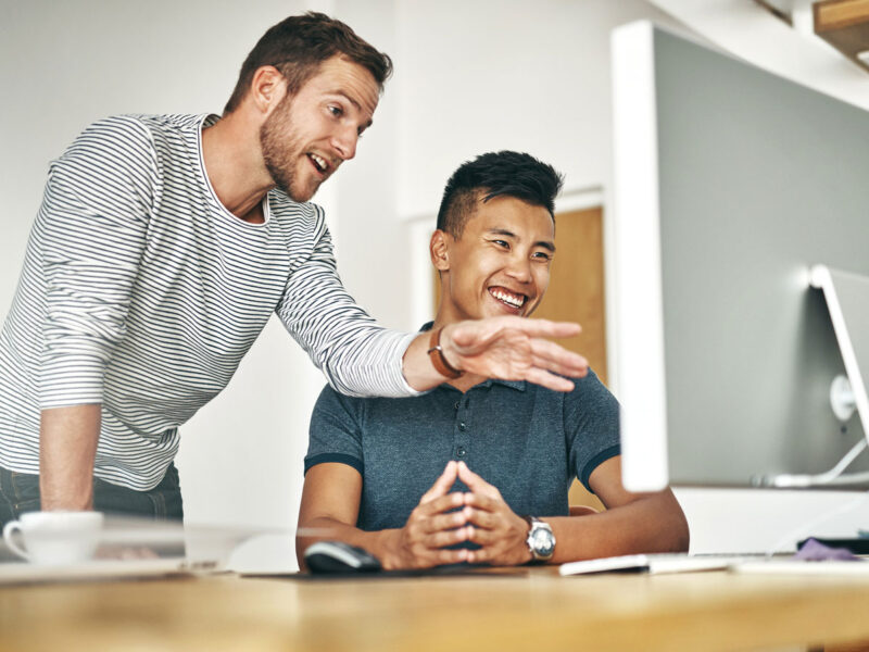 Two coworkers smiling and collaborating at a computer in an office.