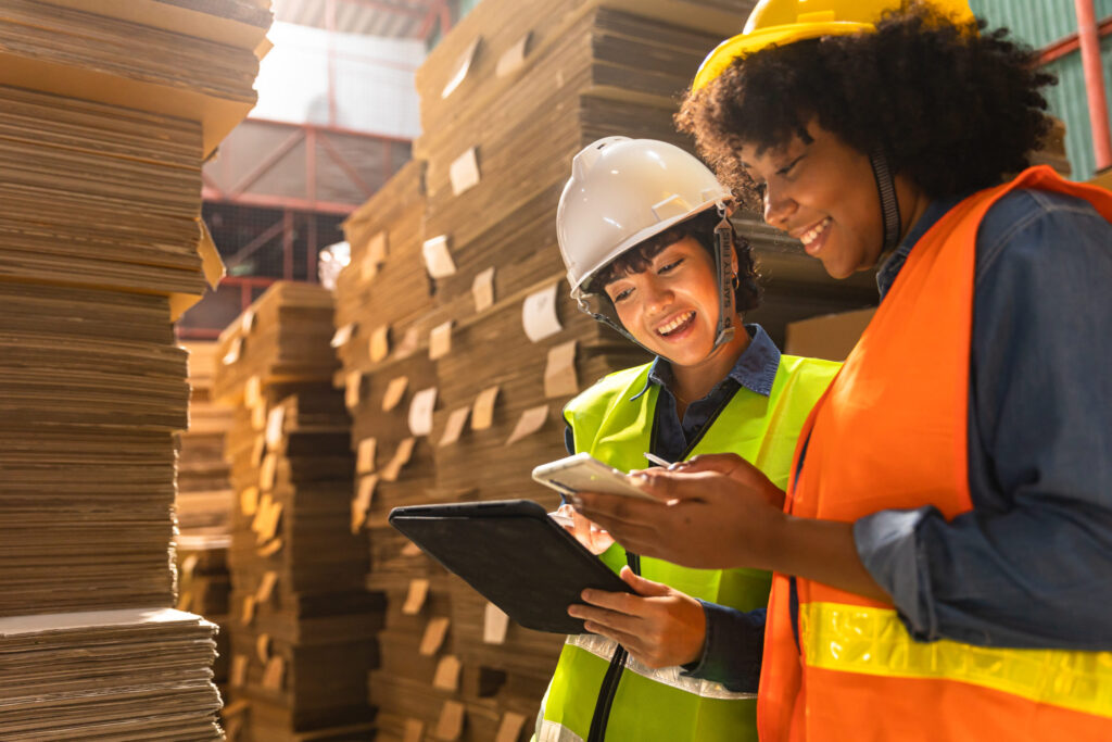 Two workers in safety vests and helmets reviewing a tablet and phone in a warehouse.