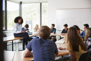 A diverse group of high school students sits around a table in a bright classroom, listening to a teacher who is perched on a desk near a whiteboard with math equations.