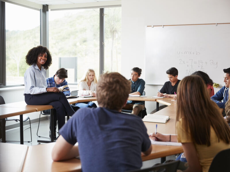 A diverse group of high school students sits around a table in a bright classroom, listening to a teacher who is perched on a desk near a whiteboard with math equations.