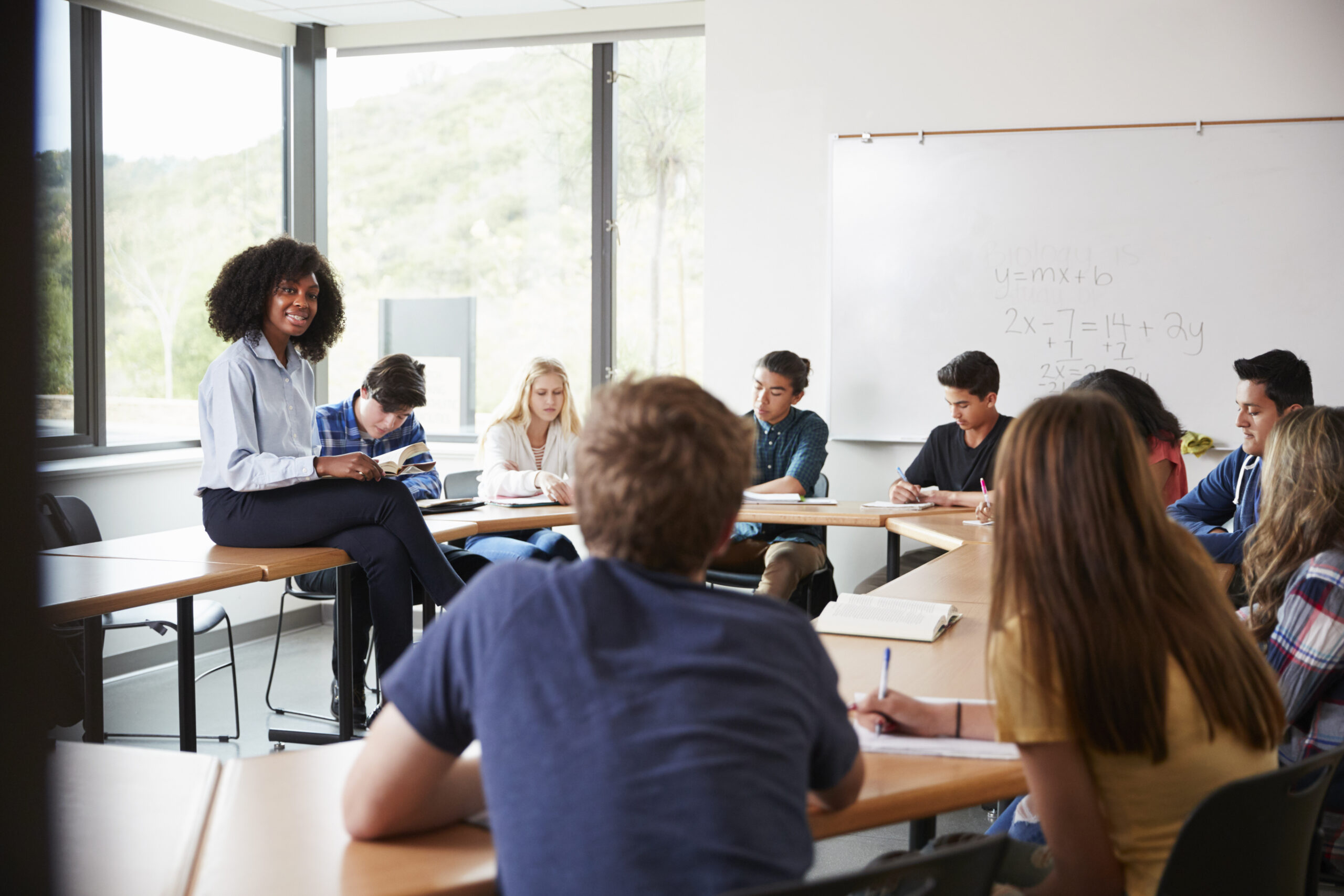 A diverse group of high school students sits around a table in a bright classroom, listening to a teacher who is perched on a desk near a whiteboard with math equations.