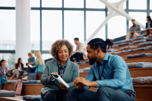 A woman in a grey blazer and a man in a blue shirt sit on wooden tiered seating, focused on a digital tablet they are holding together in a modern, sunlit atrium.