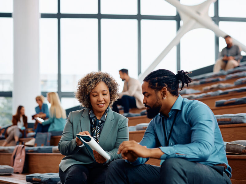 A woman in a grey blazer and a man in a blue shirt sit on wooden tiered seating, focused on a digital tablet they are holding together in a modern, sunlit atrium.