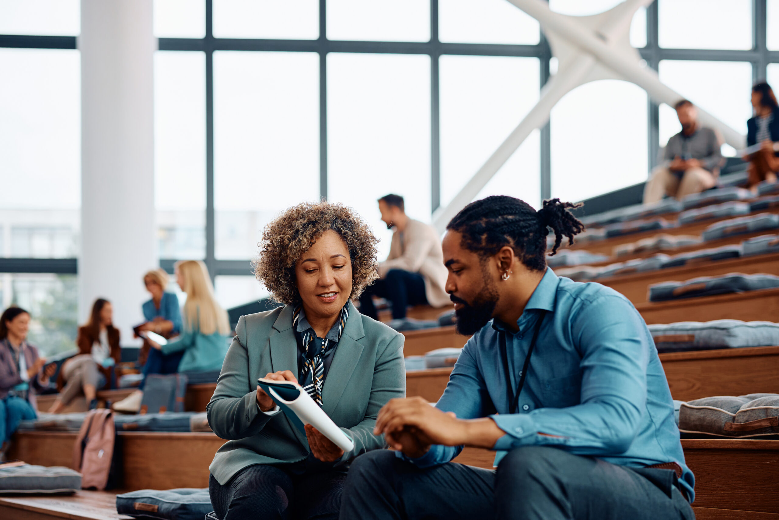 A woman in a grey blazer and a man in a blue shirt sit on wooden tiered seating, focused on a digital tablet they are holding together in a modern, sunlit atrium.
