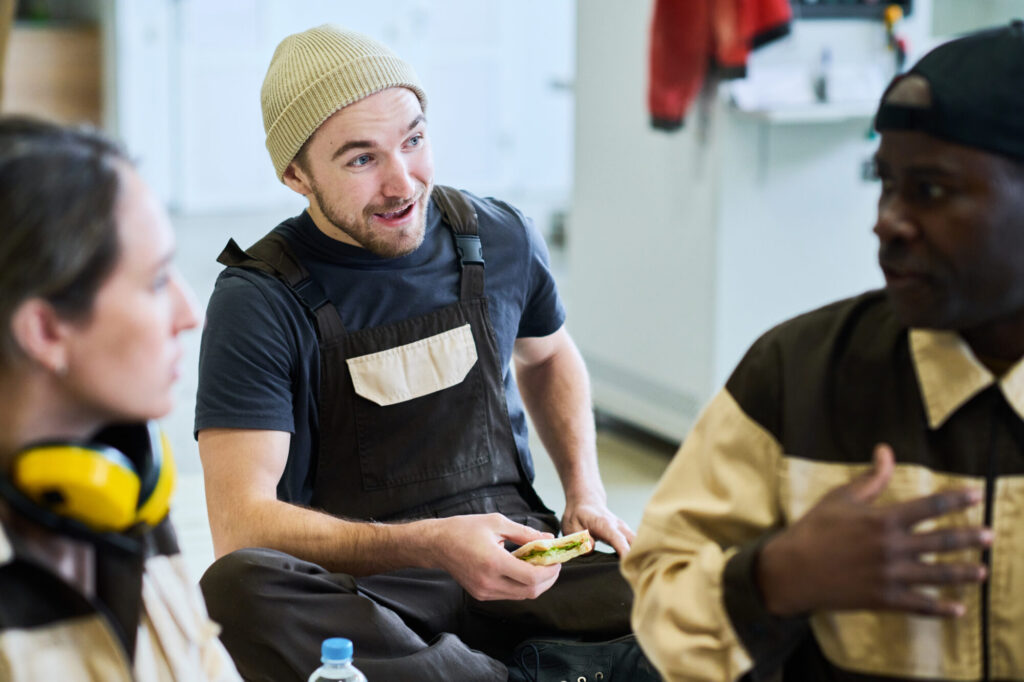 Three coworkers in work uniforms sit together on a break, chatting; one person wearing a beanie gestures while holding a sandwich.