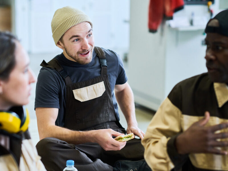 Three coworkers in work uniforms sit together on a break, chatting; one person wearing a beanie gestures while holding a sandwich.