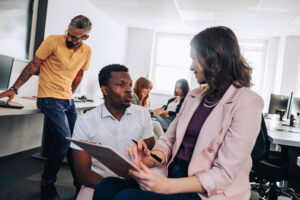 A diverse team collaborating in a bright modern office, with a woman in a pink blazer reviewing a clipboard with a seated colleague.