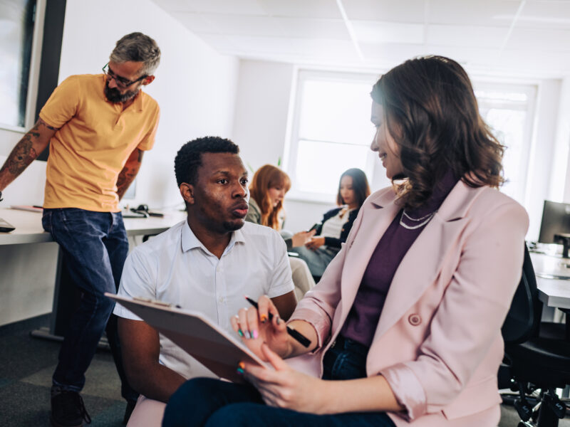 A diverse team collaborating in a bright modern office, with a woman in a pink blazer reviewing a clipboard with a seated colleague.