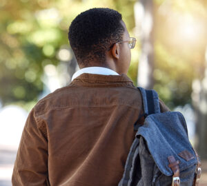A person seen from behind, wearing a brown jacket and carrying a denim backpack, walking along a sunny tree-lined path.