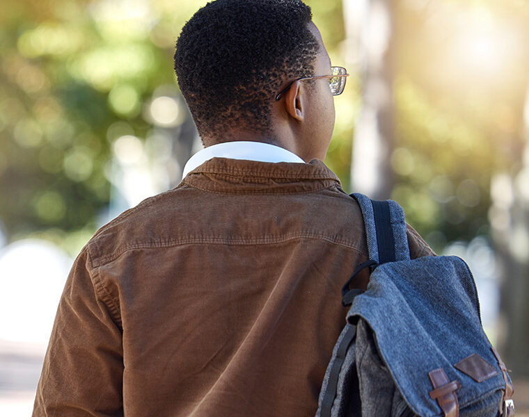 A person seen from behind, wearing a brown jacket and carrying a denim backpack, walking along a sunny tree-lined path.