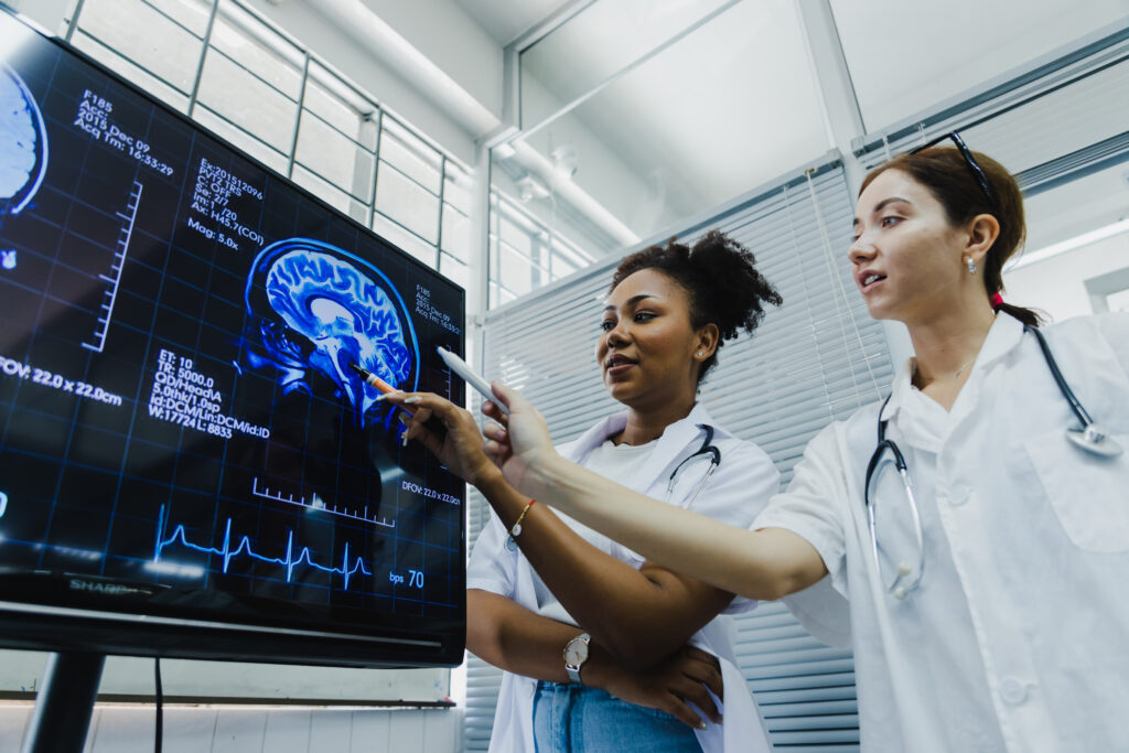 Two medical professionals in lab coats examine and discuss a digital brain scan displayed on a large monitor in a hospital setting.