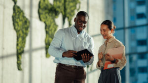 Two colleagues reviewing content on a tablet outdoors, with a green living wall world map in the background.