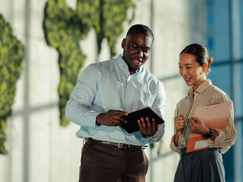 Two colleagues reviewing content on a tablet outdoors, with a green living wall world map in the background.