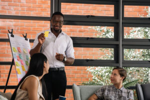 A man stands in an office setting holding a yellow sticky note, presenting to two seated colleagues next to a whiteboard filled with colorful notes.