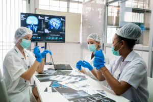 Three medical professionals wearing masks, gloves, and lab coats review brain scans and medical images together in a clinical workspace.