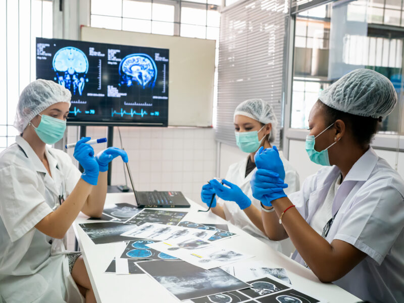 Three medical professionals wearing masks, gloves, and lab coats review brain scans and medical images together in a clinical workspace.