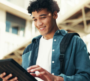 Young person smiling while using a tablet outdoors with a backpack over one shoulder.