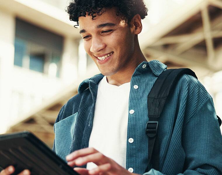 Young person smiling while using a tablet outdoors with a backpack over one shoulder.