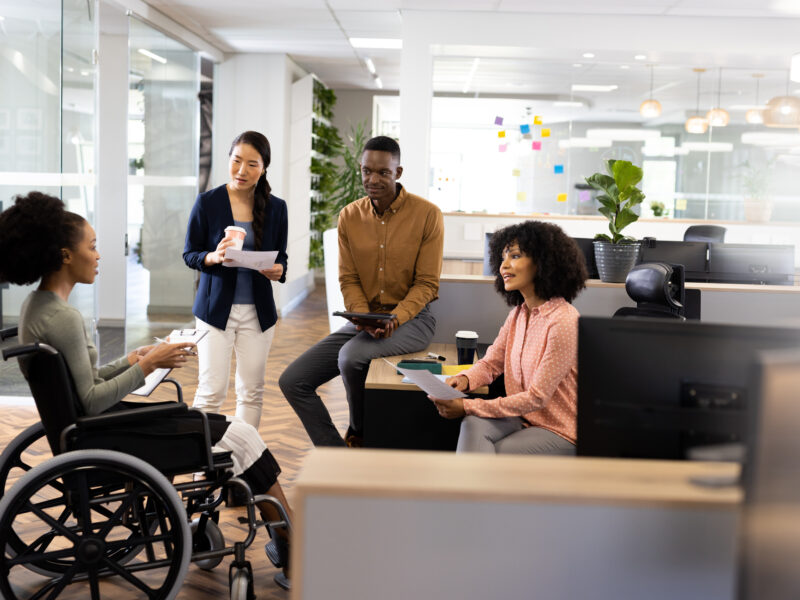 Diverse team members collaborate in a modern office, including a woman using a wheelchair, as they discuss documents and ideas together.