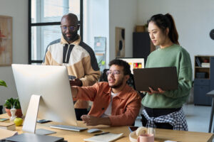 Team members collaborating around a desktop computer in a modern office, reviewing work together.