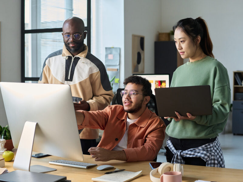 Team members collaborating around a desktop computer in a modern office, reviewing work together.