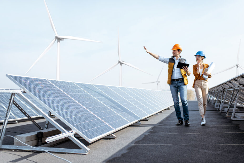 Workers inspect solar panels at a renewable energy site with wind turbines in the background.