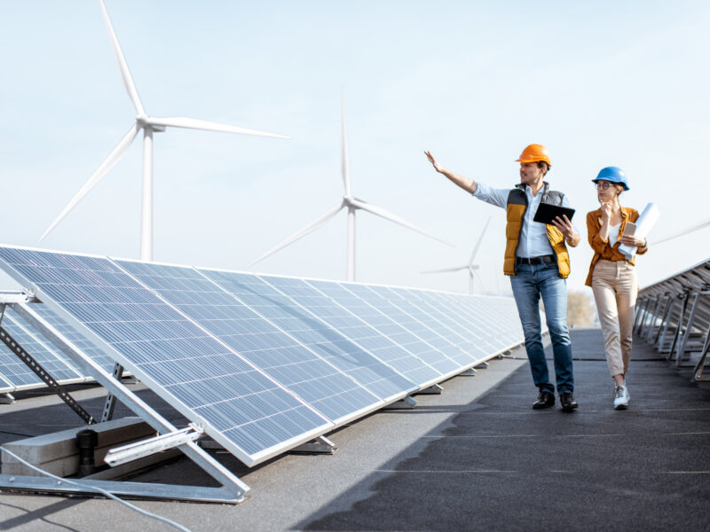 Workers inspect solar panels at a renewable energy site with wind turbines in the background.
