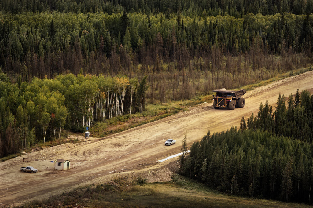 A large yellow mining haul truck travels a wide dirt road through boreal forest, dwarfing a pickup truck and small site building nearby.