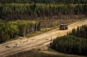 A large yellow mining haul truck travels a wide dirt road through boreal forest, dwarfing a pickup truck and small site building nearby.