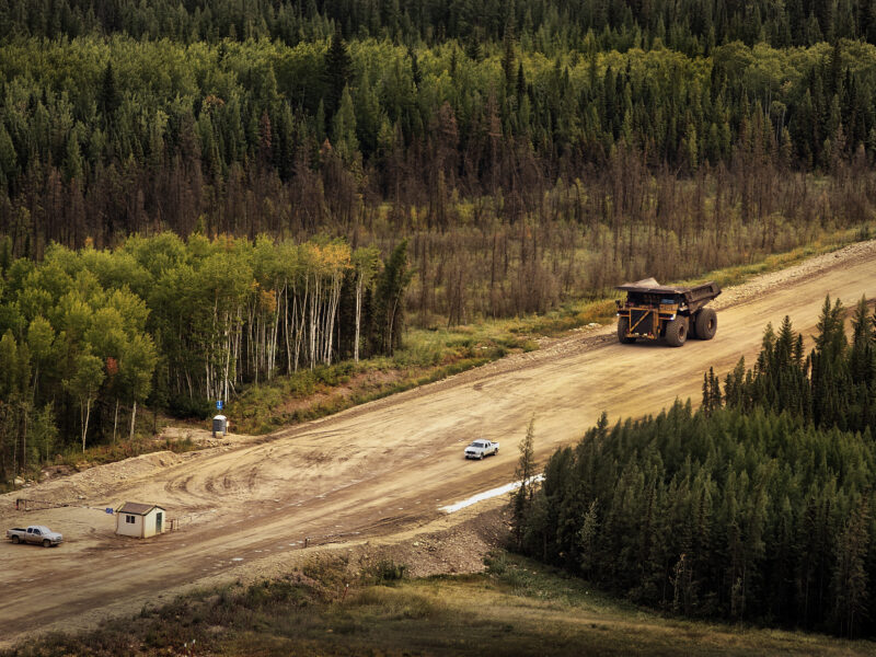 A large yellow mining haul truck travels a wide dirt road through boreal forest, dwarfing a pickup truck and small site building nearby.