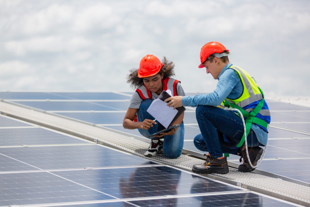 Two workers in safety gear inspect rooftop solar panels while reviewing plans and a tablet
